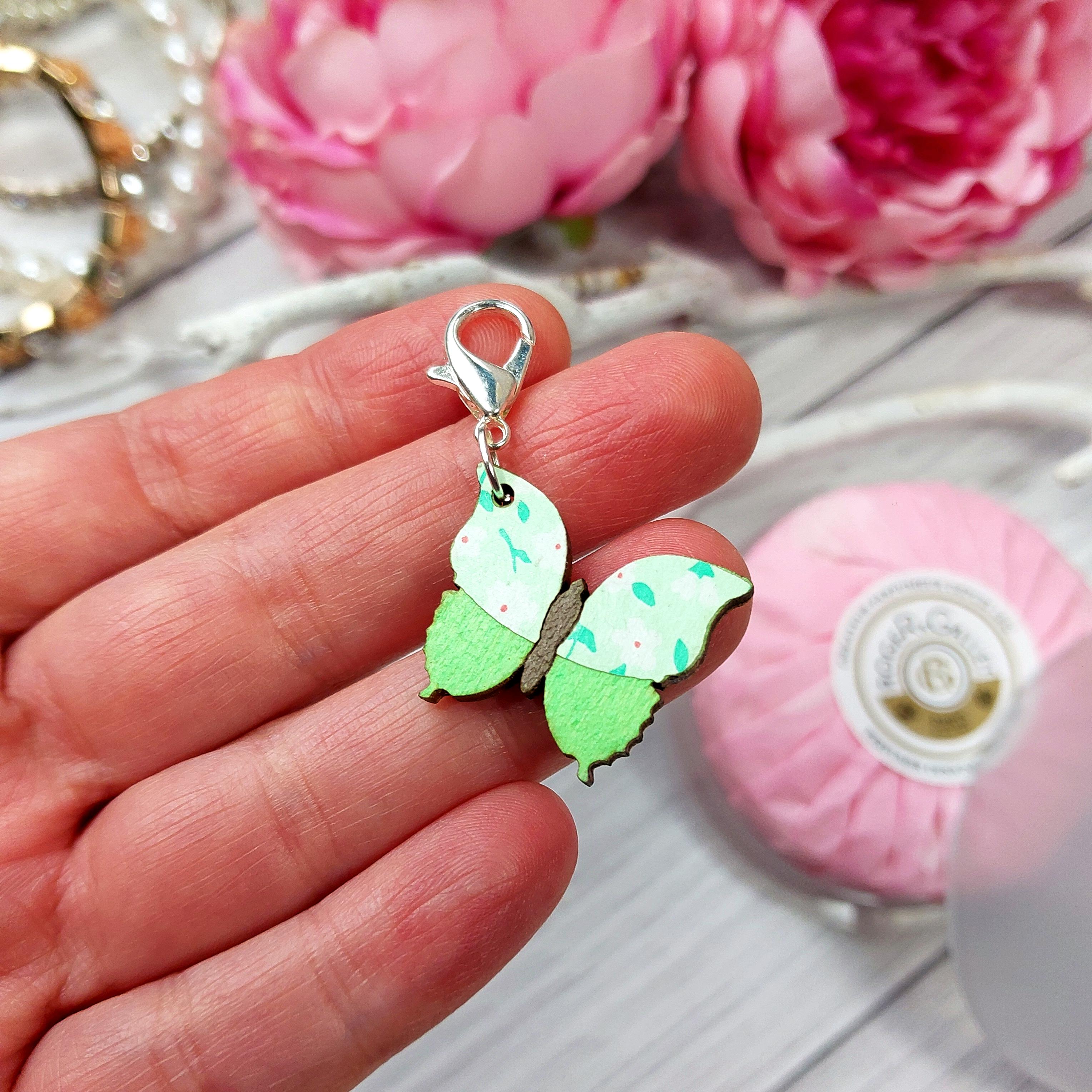 Hand holding green wooden stitch marker butterfly in front of pink flowers in the background