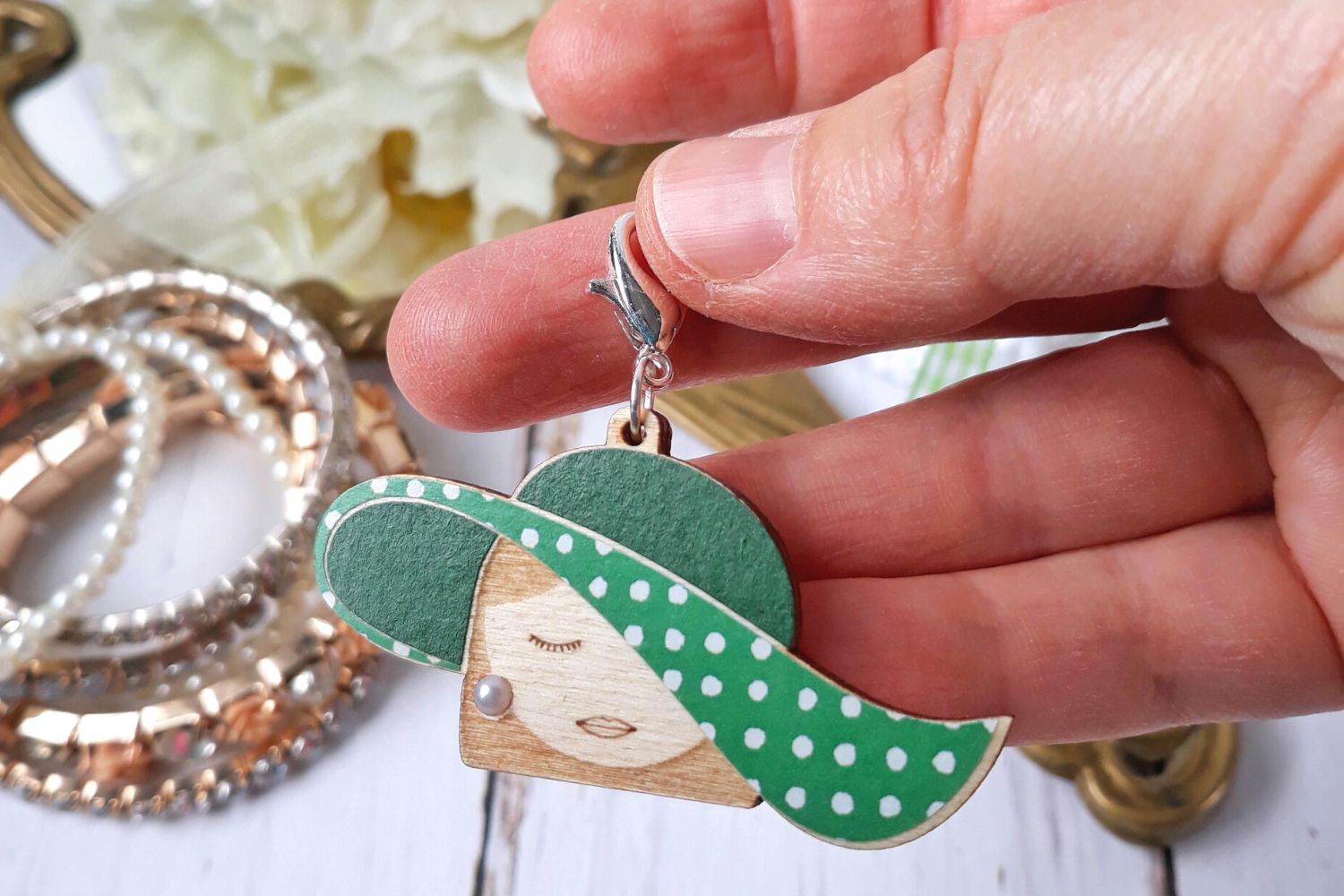 Hand holding wooden stitch marker of a lady with a green polka dot hat in front of white flowers in the background